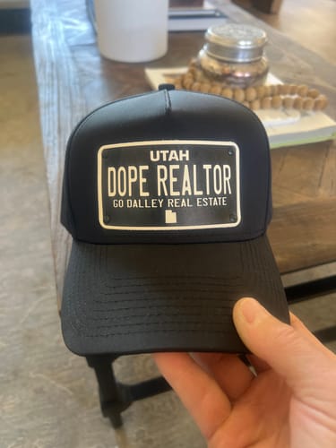 Customer holding a black Utah Blackout Plate Hat with a custom license plate patch that reads 'DOPE REALTOR'.