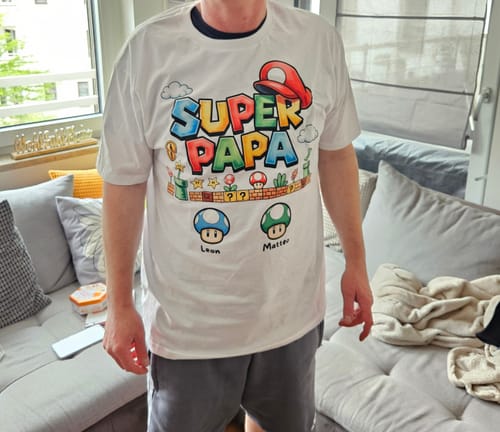 Ein Kunde trägt das personalisierte weiße "Super Papa"-T-Shirt in einem Wohnzimmer.
