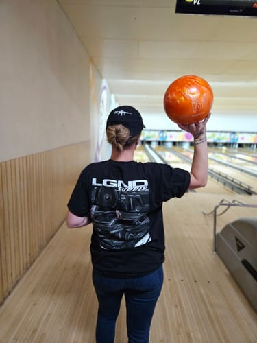 Reviewer at a bowling alley showing the back of a black t-shirt from the LGND92 Mystery CASH Box while holding a bowling ball.