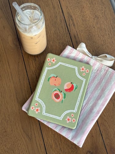 Customer's green Peachy Kindle Case with a peach design, on a pink striped cloth next to an iced coffee.