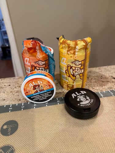 Customer's collection of different ALP nicotine pouches, including Mango Freeze and an unopened black tin, on a counter.
