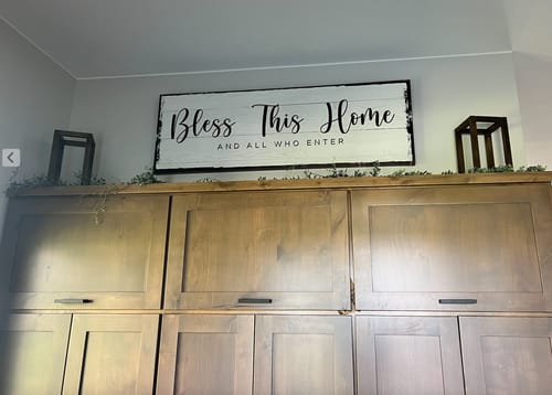 Customer's 'Bless This Home' sign with black script, displayed as wall art above tall wooden cabinets.