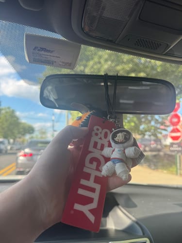 Customer holding a red air freshener and white astronaut charm hanging from a car's rearview mirror.