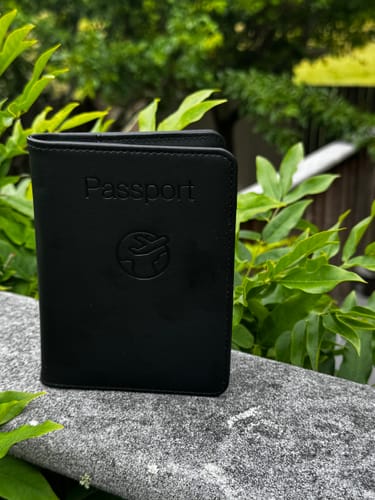 Customer's black Trackable Passport Cover standing closed on a stone ledge in front of green leaves.