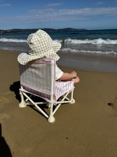 A customer's baby sits in the Billie baby chair on a sandy beach, looking out at the ocean waves.