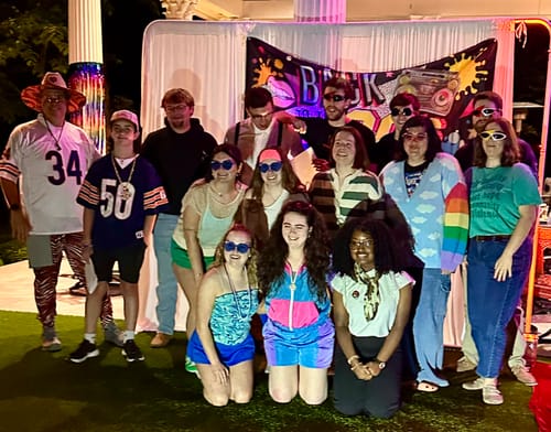 Customers in 80s costumes pose for a group photo while playing the 1980s Murder Mystery Game.