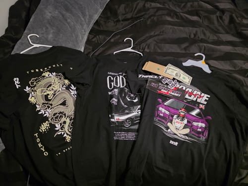 Customer's three black graphic t-shirts, sticker, and cash from the TCG94 MYSTERY CASH BOX laid out on a bed.