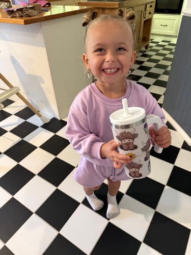 A customer's smiling child holding the Highland Cow Spill Proof Kids CREWCUP in a kitchen.