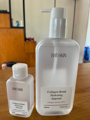 Reviewer's small 100ml and large 400ml bottles of Bad Skin Collagen Bomb Hydrating Ampoule side-by-side on a table.
