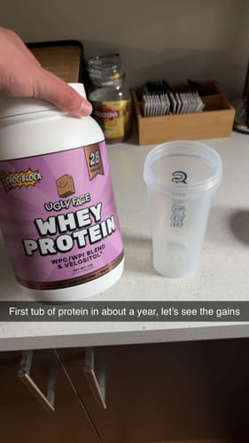 Reviewer's hand holding a tub of UGLYFACE® WHEY PROTEIN in Choc Block flavor on a counter, with a shaker cup nearby.