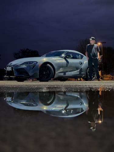 Reviewer wearing the black 80Eighty® DCG88 Shirt next to a sports car at night, reflected in a puddle.