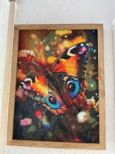 Kunde zeigt das fertige, gerahmte Diamond Painting eines bunten Schmetterlings an einer Wand.