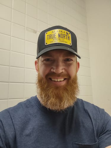 Reviewer smiling while wearing a black Alaska Plate Hat with a custom yellow license plate patch for 'True North Detailing LLC'.