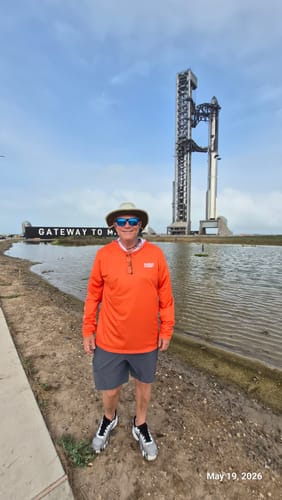 Customer wearing the orange Starship Flight 12 Sun Shirt in front of a large rocket launch tower at Starbase.