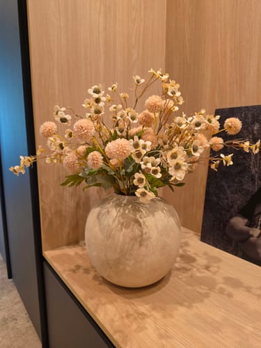 Klant toont het Wax Arrangement - Crème in een ronde, getextureerde vaas op een houten plank.