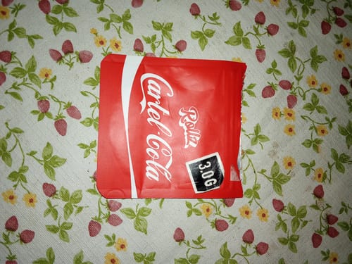 Customer's red 3.0G pouch of Cartel Cola THCA Flower resting on a floral patterned tablecloth.