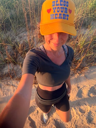 Reviewer taking a selfie outdoors wearing the yellow Bless Your Heart Trucker hat on a sand dune.