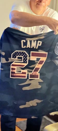 Reviewer holding the back of the custom camo baseball jersey with 'CAMP' and the number '27' in a USA flag design.