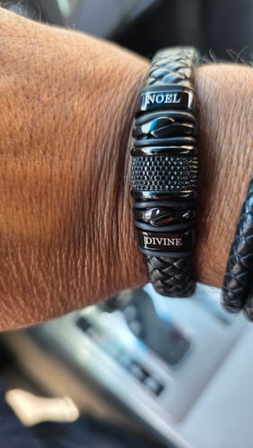 Close-up of a reviewer's wrist wearing the black braided bracelet, personalized with the names 'NOEL' and 'DIVINE'.