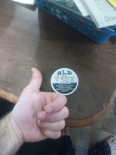 Reviewer gives a thumbs-up next to a tin of ALP Chilled Mint Nicotine Pouches on a wooden table.