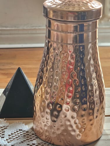 Customer's hammered copper Himalayan Ayurvedic Bedside Carafe sitting on a table next to a decorative pyramid.