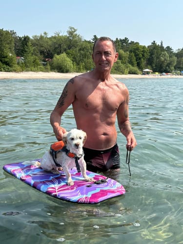 Customer with a lean, defined physique standing shirtless in the water with a small dog on a board.