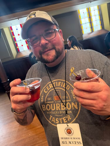 Reviewer wearing the grey Certified Bourbon Taster T-Shirt at an event, holding two drinks.