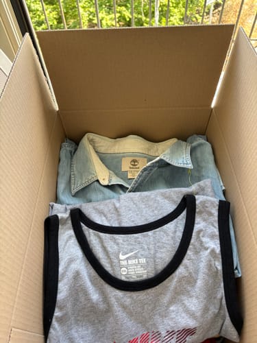 Cliente mostra il contenuto della sua box per rivendita, con in cima una camicia in denim e una canotta Nike.