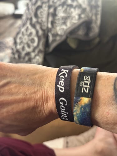 Customer's wrist wearing two fabric bracelets. One is black with 'Keep Going' in white script, the other is multicolored.