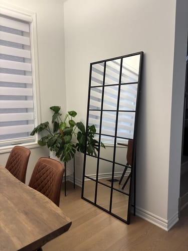 Customer's modern black grid wall mirror leaning against a wall in a dining room next to a plant.