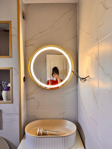 Oglinda LED rotundă AURA a clientului, cu ramă aurie, montată într-o baie modernă, cu lumina LED aprinsă.