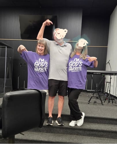 Two reviewers in purple God's Favorite t-shirts, playfully pointing at a person standing between them on a stage.