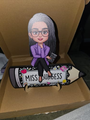 Customer's personalized wooden plaque of a teacher figure sitting on a pencil printed with 'Miss Burgess', shown on its stand.