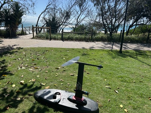Customer's hydrofoil board with the Foil Drive Assist MAX system installed, resting on grass near a beach.