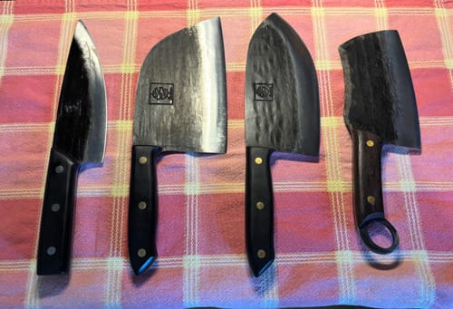 Customer's four varied Mystery Knives with dark, forged blades and black handles on a tablecloth.