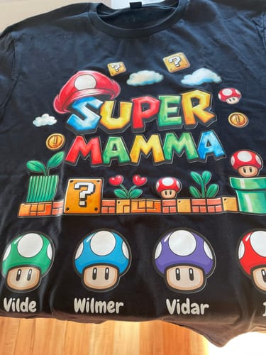 Rezensent zeigt das personalisierte schwarze „Super Mamma“-T-Shirt mit einem Design im Videospiel-Stil und den Namen der Kinder.