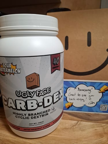 Reviewer's white tub of UGLYFACE CARB-DEX in Sour Watermelon flavor on a wooden surface with a handwritten note.