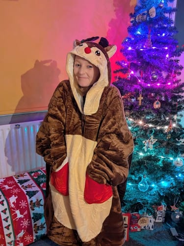 Customer wearing the soft, oversized Reindeer Wearable Blanket Hoodie with hands in the red mitten pockets by a Christmas tree.