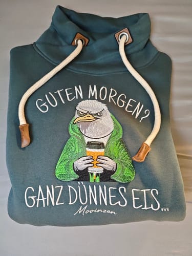 Kunde zeigt das gefaltete grüne Sweatshirt mit der hochwertigen Stickerei einer mürrischen Möwe mit Kaffeebecher.