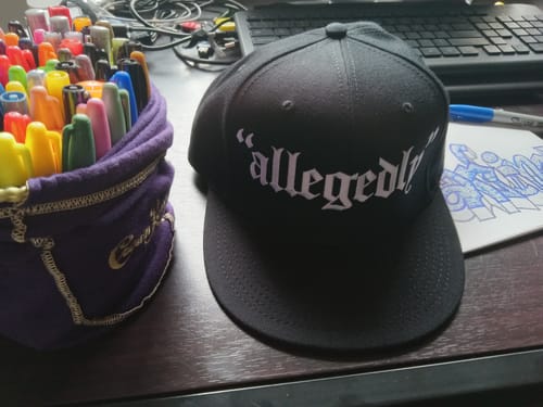 Customer's black 'Allegedly' Snapback sitting on a desk next to art supplies.