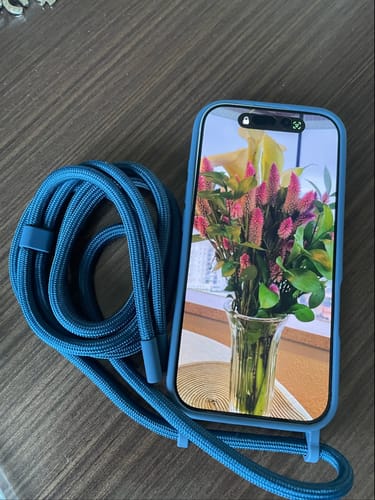 A case 'Azul da Cor do Mar' de um cliente em um celular, com a alça de corda azul enrolada ao lado em uma mesa de madeira.