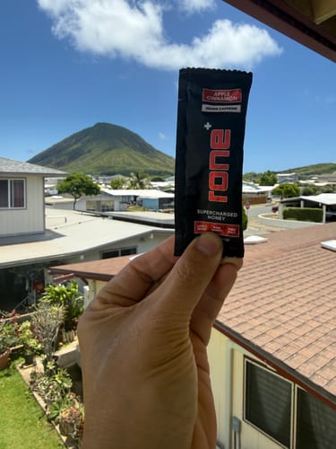 Customer's hand holding a single packet of Apple Cinnamon SuperCharged Honey with a scenic mountain view in the background.
