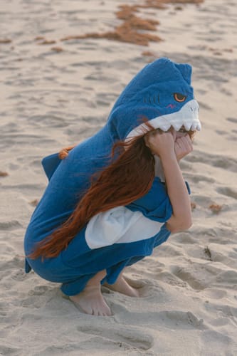 Reviewer crouching on a sandy beach wearing the blue shark surf poncho, showing the detailed hood.