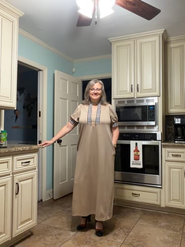 Reviewer standing in a kitchen wearing the flowy, beige Timeless Plaid Trim Maxi Dress.