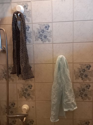 Vásárló két Silkura hámlasztó törölközője, egy sötét és egy világoszöld, fürdőszobai csempén lógva.