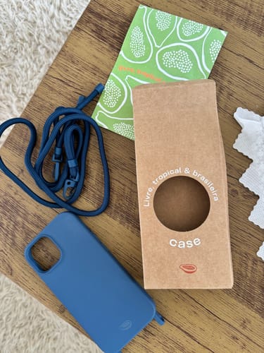 Foto de cliente da Cordinha Nora azul, exibida com uma capa de celular e a embalagem do produto sobre uma mesa.