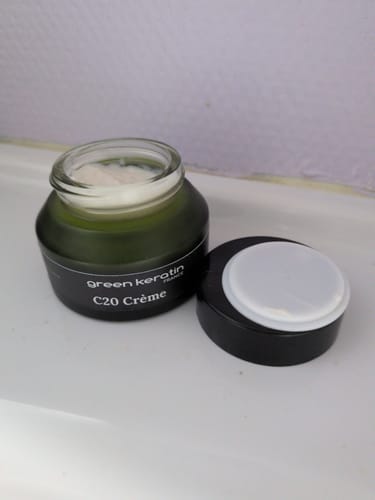 Photo d'un client montrant le pot vert ouvert de C20 Crème pour le visage sur une surface blanche, avec le couvercle à côté.