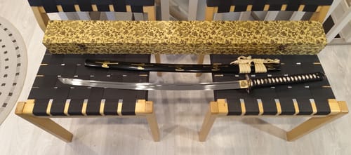 Customer's Custom Japanese Sword displayed unsheathed on a bench, with its black scabbard and decorative box.