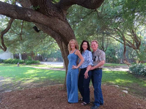 Reviewer in the Evolet Denim Jumpsuit posing with two people under a large tree.