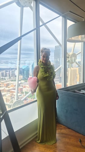 Reviewer wearing the green Naked Ruffle Halter Maxi Dress, standing in front of a window with a city view.